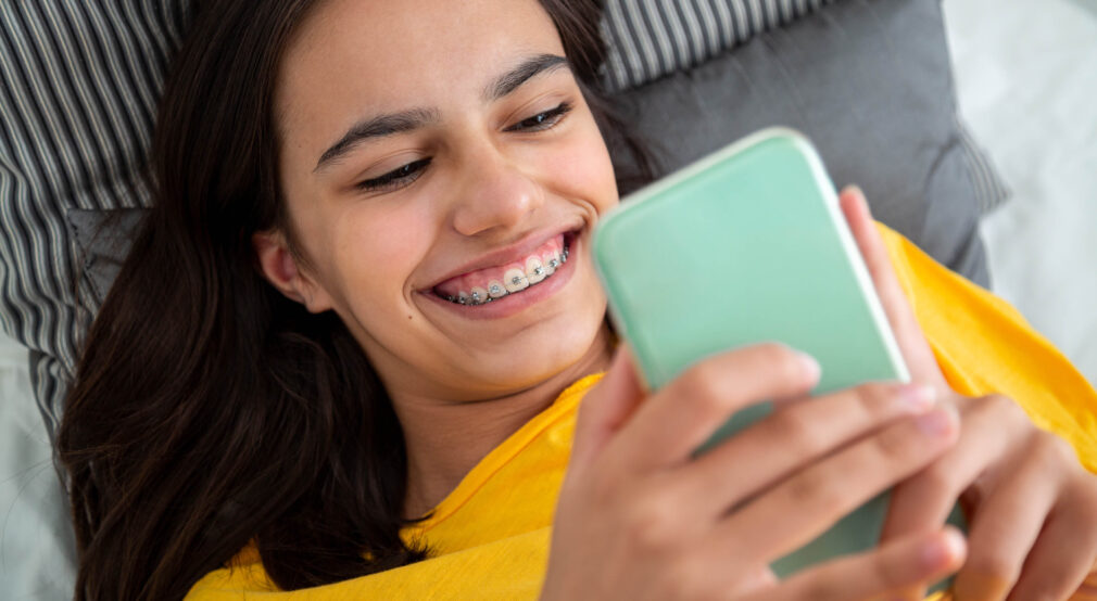 young teen with braces in yellow shirt, laying on a couch, looking at a cell phone