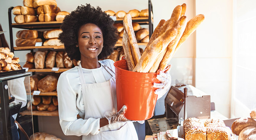 Cheerful female baker is standing and holding a basket of pastry. She is proposing food to her customer. The African-American woman is looking forward and smiling. Realizing a dream