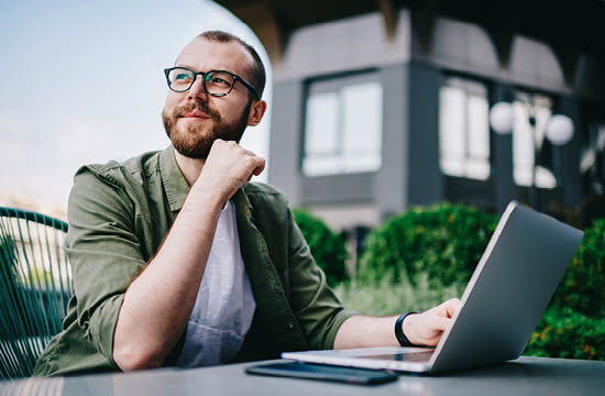 Caucasian man with glasses in a white shirt and green button down sits in front of a laptop outside