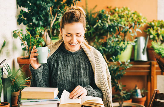woman in green sweater with cup of coffee. surrounded by plants and books.