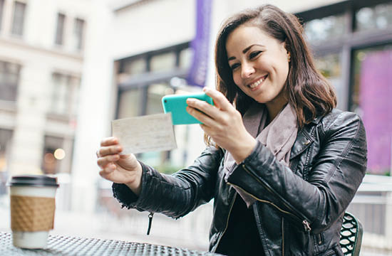A beautiful Caucasian adult woman sits in a New York city park, taking a picture of a check with her smart phone for a Remote Deposit Capture. She smiles, wearing modern stylish clothing with darker and black colors.
