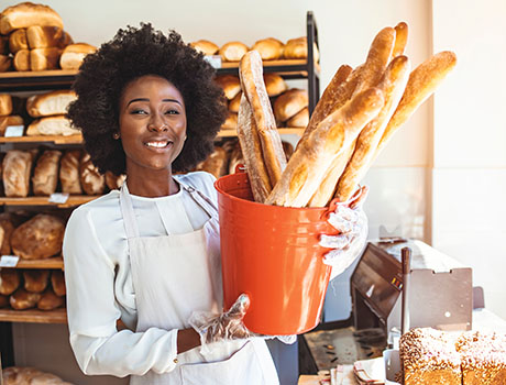 Cheerful female baker is standing and holding a basket of pastry. She is proposing food to her customer. The African-American woman is looking forward and smiling. Realizing a dream. Small business concept