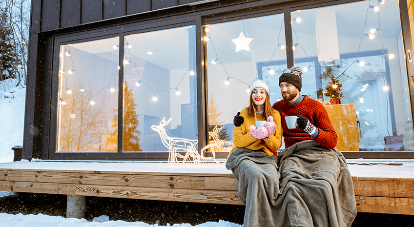 couple holding mugs and sitting on their porch in the winter with snow on the ground, wearing sweaters, beanie hats, gloves, and a blanket